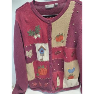 May include: Burgundy button-up cardigan with embroidered autumn-themed patches. Features pumpkins, leaves, acorns, and a birdhouse design. The sweater has long sleeves and a round neckline.