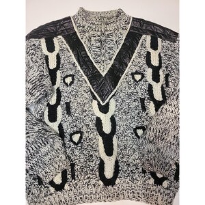 May include: A black, white, and gray knit sweater with a high collar and black leather accents. The sweater features a V-neck design and decorative black and white patterns. The sleeves are long and the overall style is vintage.