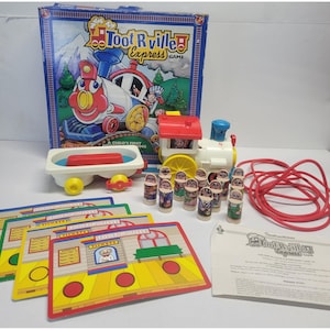 May include: A vintage Toot R Ville Express board game with a train-themed design. The game includes a box, train engine, train car, game pieces, cards, and instructions. The box features a colorful illustration of a train and mountains.