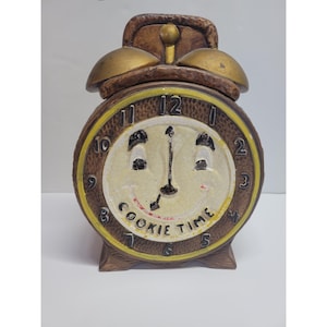 Puede incluir: Un tarro de galletas de cerámica marrón y dorada con forma de despertador. La esfera del reloj tiene una cara sonriente y las palabras "COOKIE TIME". El tarro tiene una superficie texturizada y una tapa con dos campanas doradas.