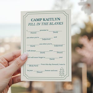 May include: A cream-colored fill-in-the-blanks card with green text that reads "Camp Kaitlyn." The card has a decorative border and prompts for a name, verb, adjective, and noun. The card is held in a hand.