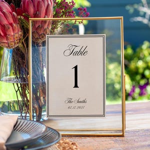 以下が含まれることがあります： 「Table」の文字と黒い数字「1」が入った、金色のフレームのテーブルナンバーカード。カードには「The Smiths」という名前と日付「07.17.2027」も含まれています。背景には花瓶に入った花があります。