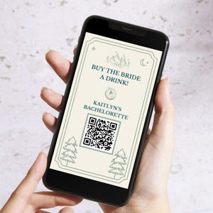 May include: A black smartphone displays a digital invitation with the text "BUY THE BRIDE A DRINK!" and "KAITLYN'S BACHELORETTE." The invitation features a QR code, mountain and tree graphics, and a decorative border.
