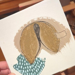 Puede incluir: Un lienzo pintado a mano con una galleta de la fortuna con un diseño marrón y negro. La galleta está pintada sobre un fondo blanco con un patrón de lunares verdes y blancos.