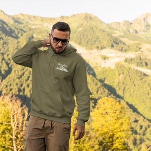 Puede incluir: Una sudadera con capucha verde oliva con un gráfico de montaña blanco y la palabra "Mountain" impresa encima. La sudadera tiene un bolsillo delantero y una capucha con cordón. La persona lleva gafas de sol y pantalones marrones. El fondo es una cordillera.