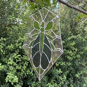 May include: Stained glass insect suncatcher in shades of green and clear glass. The insect design features a detailed body and wings, with a geometric shape. The suncatcher is hanging from a silver chain, perfect for a garden or window display.