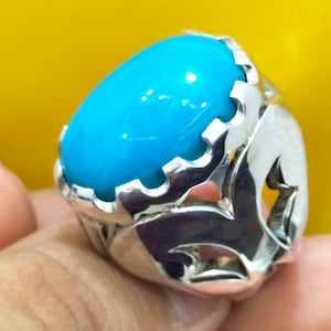 May include: A silver ring featuring a large, oval-shaped, bright turquoise stone. The ring's setting has a decorative, openwork design with a polished finish. The ring is shown against a yellow background.