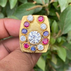 Puede incluir: Un anillo dorado con una cara rectangular, adornado con una gran piedra preciosa transparente central rodeada de piedras más pequeñas y coloridas. El anillo presenta un halo de piedras rojas, azules y transparentes alternas, creando un contraste llamativo.