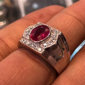Könnte beinhalten: Ein silberner Ring mit einem großen ovalen Rubin, der von kleineren weißen Edelsteinen umgeben ist.