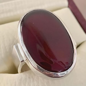 May include: A silver ring featuring a large, oval-shaped, deep burgundy stone. The stone is set in a polished silver band with a simple, modern design. The ring's setting is smooth and the stone has a glossy finish.