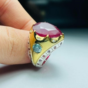Puede incluir: Un anillo de plata y oro con una gran piedra roja rodeada de piedras rojas y blancas más pequeñas. El anillo tiene un diseño decorativo con una piedra azul en el lateral.