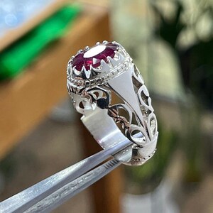 Peut inclure: Une bague en argent avec un motif orné et une grande pierre précieuse rouge foncé de forme ovale.