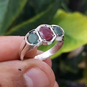 May include: A silver ring featuring three oval-shaped gemstones. The central stone is a deep red ruby, with two flanking green emeralds.