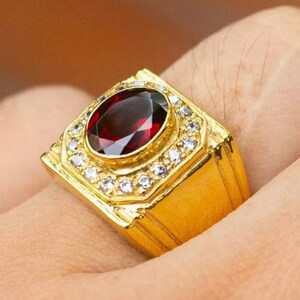 May include: A gold ring with a large red oval gemstone surrounded by smaller white gemstones.