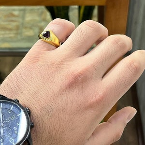 May include: A gold ring with a triangular red gemstone. The ring is on a person's finger.