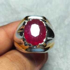 Könnte beinhalten: Ein silber- und goldfarbener Ring mit einem großen, oval geschliffenen Rubin. Der Rubin hat eine tiefrote Farbe und ist der Mittelpunkt des Rings. Der Ring hat eine polierte Oberfläche und ein klassisches Design.