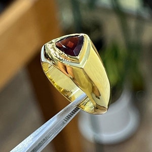 May include: A gold ring with a triangular red gemstone set in the center. The ring has a thick band and a smooth, polished finish.