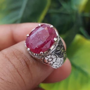 May include: A silver ring featuring a large, oval ruby gemstone. The ring band is adorned with intricate floral designs.