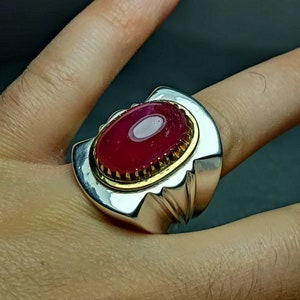 May include: A silver ring with a large, oval-shaped, red gemstone set in a gold-toned bezel. The ring has a detailed, textured band.