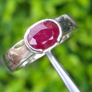 May include: A silver ring featuring a large, oval-cut ruby gemstone. The ruby is a deep, vibrant red color and is set in a simple, polished silver band. The ring is shown against a blurred green background, highlighting the gemstone's brilliance.