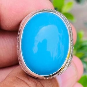 Peut inclure: Une bague de forme ovale avec une pierre turquoise lisse et brillante sertie sur une bande en métal argenté. La monture métallique présente un motif texturé détaillé sur le bord extérieur, encadrant la pierre bleue vive polie.