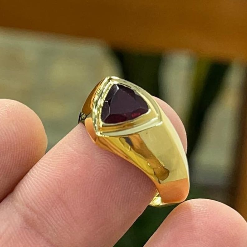 May include: A gold ring with a triangular red gemstone set in the center. The ring has a thick band and a polished finish.