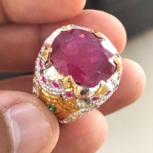 May include: A silver and gold ring with a large, oval-shaped ruby gemstone. The ring is set with smaller rubies and diamonds around the main stone.