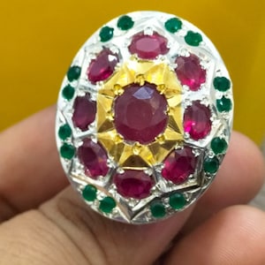 May include: A silver ring with a large red gemstone surrounded by smaller red gemstones and green gemstones.