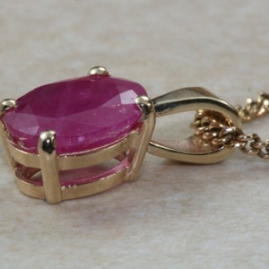 May include: A gold pendant necklace featuring a large oval ruby gemstone set in a gold frame. The ruby is a deep pink colour and has a high polish.
