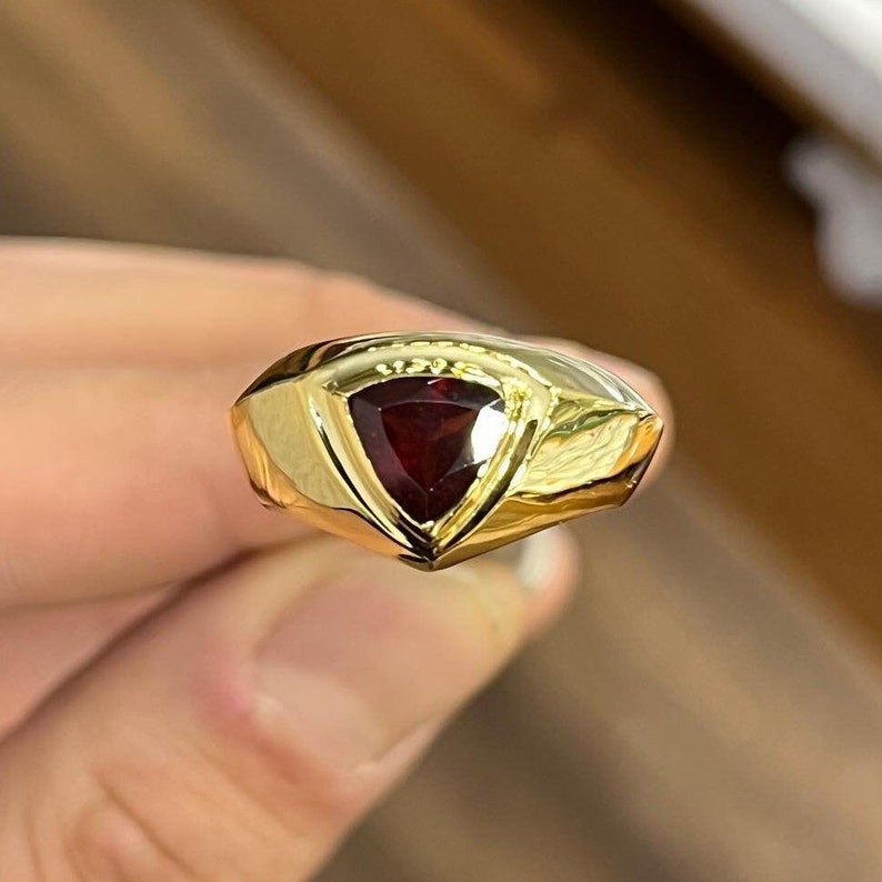 May include: A gold ring with a triangular red gemstone set in the center. The ring has a smooth, polished finish.
