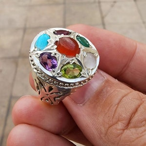 Op de afbeelding: Een zilveren ring met een rond ontwerp met zeven edelstenen: turquoise, rood, groen, wit, paars, groen en rood. De ring heeft een gedetailleerd, sierlijk ontwerp.