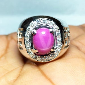 Puede incluir: Un anillo de plata con una piedra preciosa de zafiro estrellado rosa rodeada por un halo de piedras blancas más pequeñas.