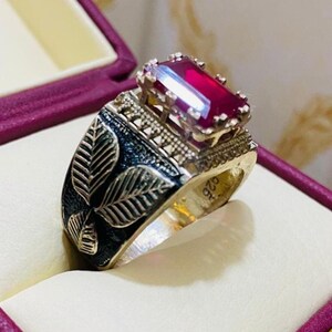 May include: A silver ring with a large, rectangular, red gemstone. The ring has a detailed design with black leaf patterns on the band and intricate metalwork around the gem. The ring is displayed in a purple jewelry box.