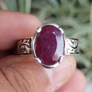 May include: A silver ring featuring an oval-cut, deep red ruby gemstone. The ring's band has an engraved floral design on the sides. The ruby is set in a silver bezel setting, showcasing its rich color and clarity.