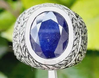 May include: A silver ring with an oval, deep blue gemstone. The gemstone is faceted, catching the light. The ring's band has a detailed, woven pattern around the gemstone, adding texture and visual interest. The ring is held by a silver tool.