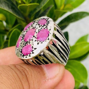 May include: Silver ring featuring oval-cut pink gemstones. The ring has a detailed design with black stripes on the sides and a decorative setting around the gemstones. The gemstones are arranged in a cluster.