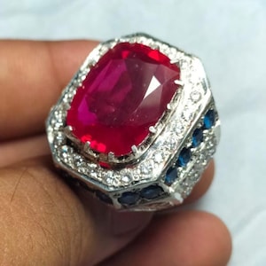 Könnte beinhalten: Ein silberner Ring mit einem großen, rechteckigen Rubin. Der Rubin ist tiefrot und von kleineren, klaren Edelsteinen umgeben. Der Ring hat kleine blaue Edelsteine an den Seiten.