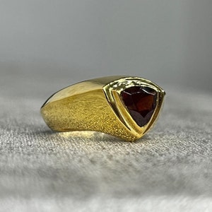 May include: A gold ring with a triangular red gemstone set in the center. The ring has a smooth, curved band.