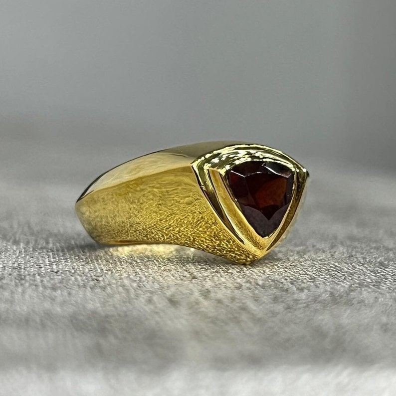 May include: A gold ring with a triangular red gemstone set in the center. The ring has a smooth, curved band.