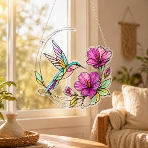 May include: A circular stained glass suncatcher featuring a colorful hummingbird in flight alongside vibrant pink hibiscus flowers and green leaves. The design is outlined in black and suspended by a silver metal chain against a bright window.