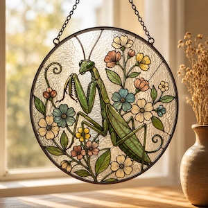 May include: A circular stained glass suncatcher featuring a green praying mantis surrounded by various colorful flowers including blue, yellow, and pink blossoms with green leaves, all encased in a dark metal frame with a hanging chain.
