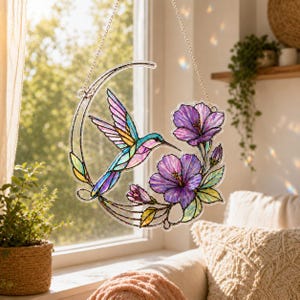 May include: A crescent-shaped suncatcher featuring a stained glass style design of a hummingbird in flight with iridescent blue, pink, and yellow wings, positioned next to two large purple hibiscus flowers and green leaves, suspended by a silver chain.