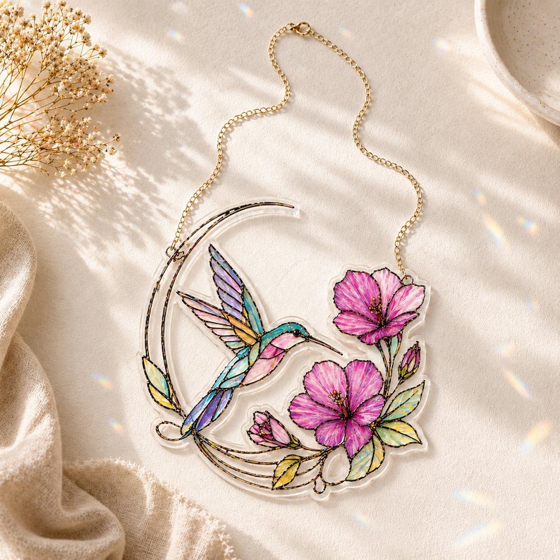 May include: A decorative hanging ornament featuring a clear acrylic crescent moon shape with a colorful hummingbird and two vibrant pink hibiscus flowers. The design includes green leaves and a gold-toned metal chain for hanging the piece.