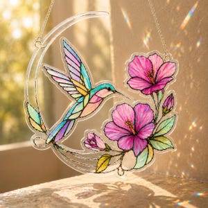 May include: A clear acrylic suncatcher featuring a colorful hummingbird in flight alongside vibrant pink hibiscus flowers and green leaves, all arranged in a crescent moon shape. The design includes black outlines and hangs from a silver chain.