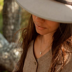 May include: A gold necklace featuring a small, white, floral pendant. The necklace is worn with a button-down shirt and a wide-brimmed hat. The pendant is a detailed, delicate design.