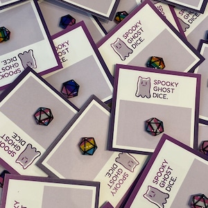 May include: Several polyhedral dice with a rainbow gradient, displayed on cards that read "Spooky Ghost Dice." The dice feature a geometric design with black borders. The cards are light purple with a white rectangle and a ghost illustration.