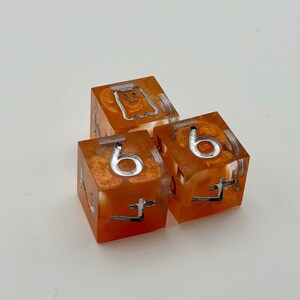 May include: Three orange resin dice with silver numbering and symbols. Each die features a six and a unique symbol. The dice are set against a white background.