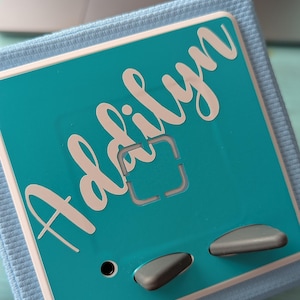 May include: A teal blue square box with a white name "Adalyn" written in cursive. The box has two gray buttons and a black audio jack.