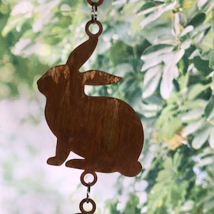 May include: A rusty metal rabbit silhouette hanging from a chain. The rabbit is facing to the right and has a long, pointed ear. The background is blurred and shows green foliage.