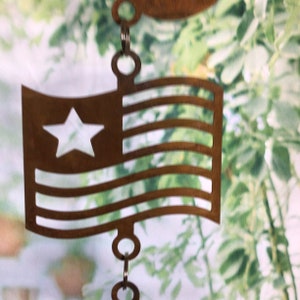 May include: A metal wind chime featuring a cut-out design of an American flag with a white star and red and white stripes. The chime is hanging from a chain with multiple rings.